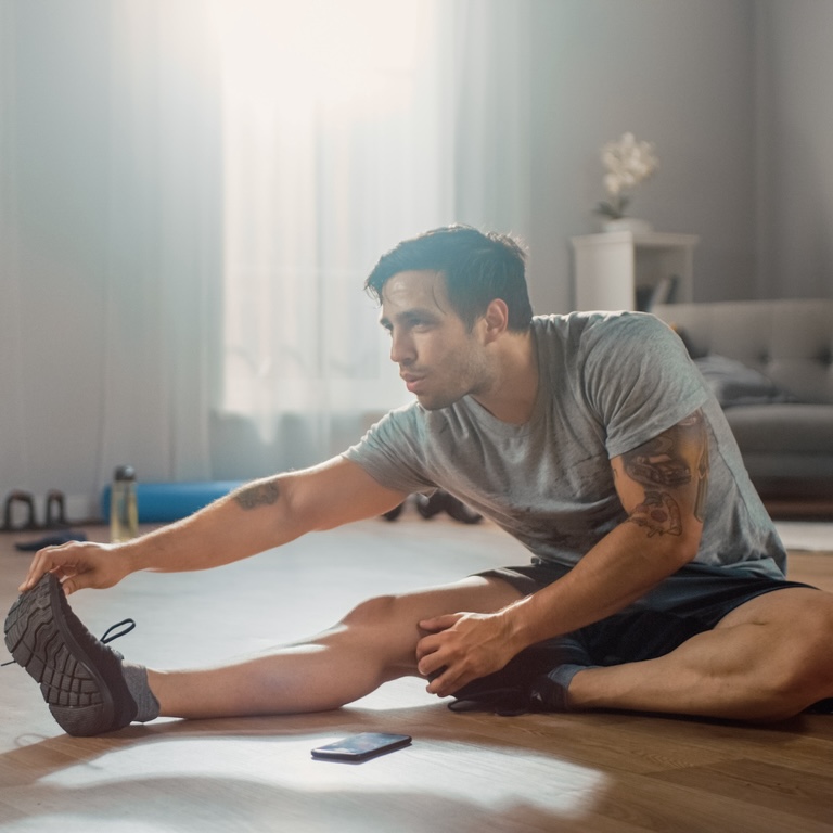 Strong Athletic Fit Man in T-shirt and Shorts Stretches His Body for Morning Exercises at Home in His Spacious and Bright Living Room with Minimalistic Interior.